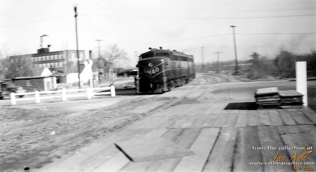Brakeman rides GM&O Alco across Hwy 79
A brakeman rides the Alco, presumably on its way back from the CB&Q yard with some cars.  Note the old dumptruck waiting at the crossing to left.  The large building at far-left is the Wells Lamont glove factory.
